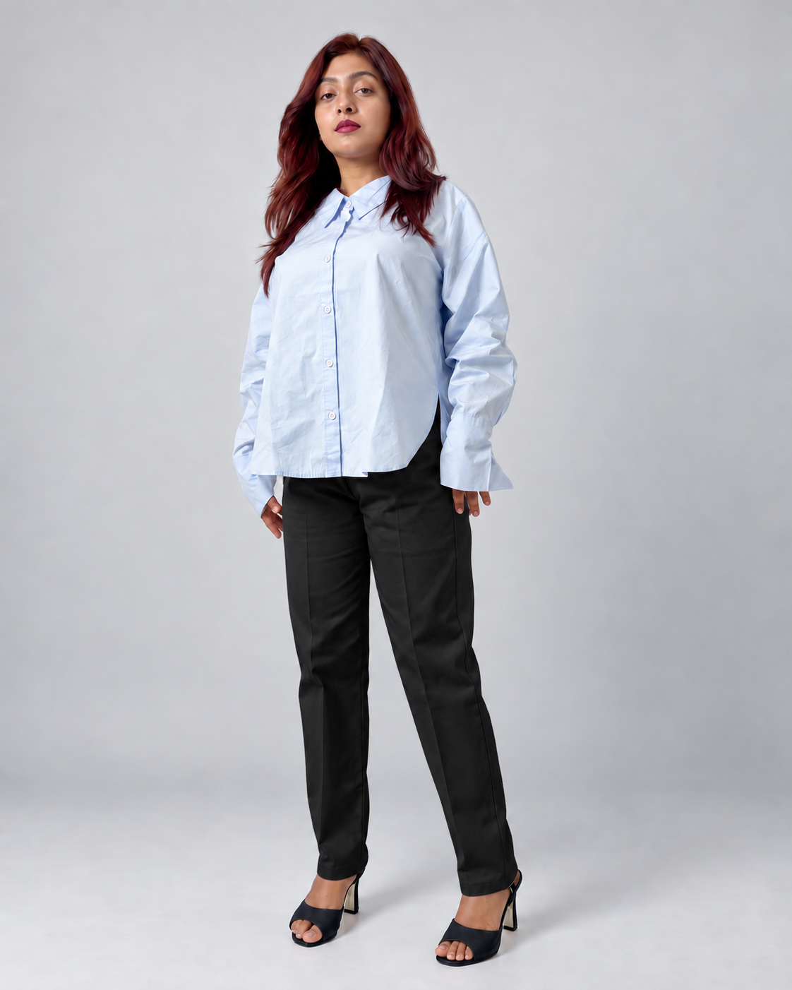 A woman modeling a light blue long-sleeve button-up shirt and black trousers against a plain light gray studio background.