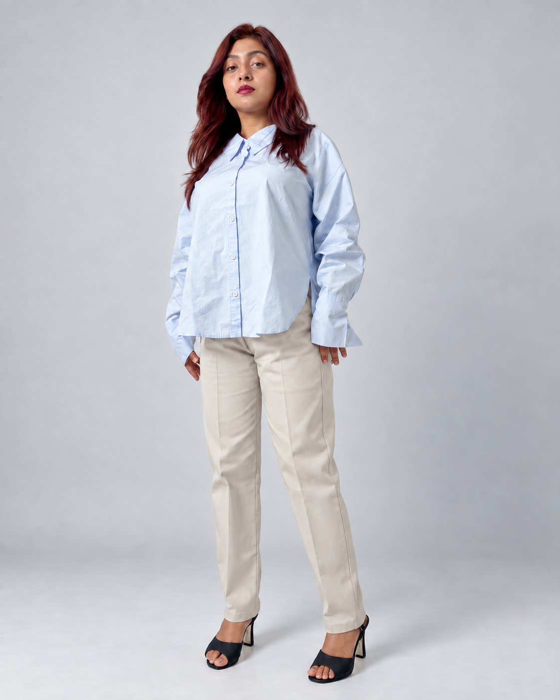 A woman modeling a light blue long-sleeve button-up shirt and beige straight-leg trousers with black heeled sandals, standing against a light gray studio background.